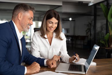 Two professionals smiling and collaborating at a laptop in an office setting.