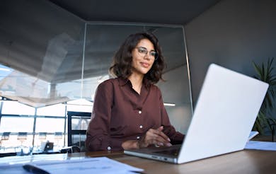 Professional woman in a brown blouse working on a laptop at a modern office desk.