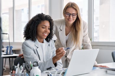 Two professional women collaborating at a desk, smiling while looking at a laptop screen.