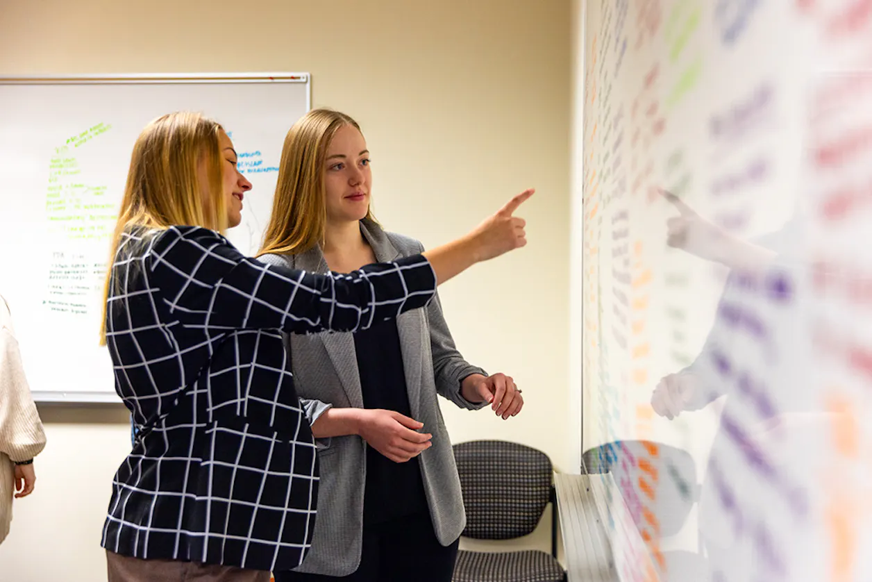 Two college students in conversation at a classroom table, one pointing at a whiteboard filled with colorful handwritten notes and brainstorming ideas.