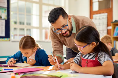 An elementary school teacher bends down to assist two young students with their classwork during a writing activity in a bright classroom.