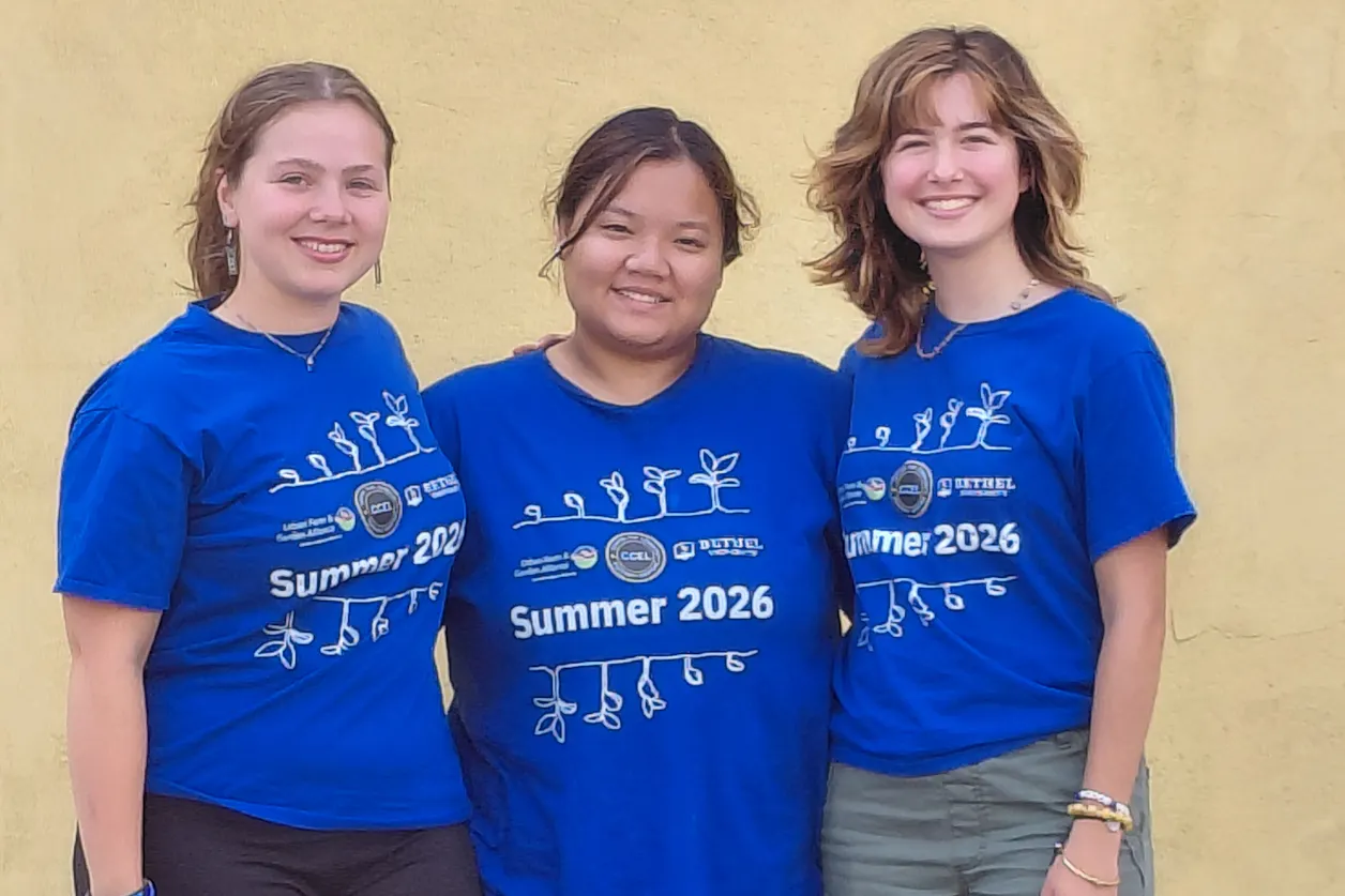 Three young women, Anna Pankratz (Social Work) - Left, Htee Wah Moo (International Relations & History) - Middle, and Zoe Diaz (Art) - Right, are wearing bright blue t-shirts with white imprint of plants growing, the words Summer 2026, and several logos for the sponsoring organization.