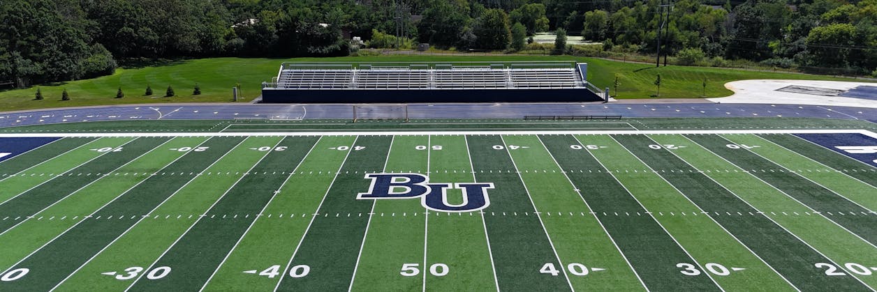 An outdoor view with the Bethel University football field in the foreground, circled by the track, with bleachers for fans in the distance.
