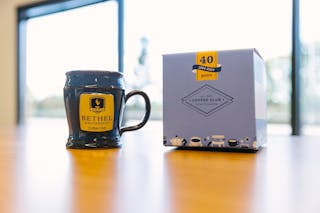 Blue pottery coffee cup with a handle that haas the Bethel University logo with the words "Coffee club" underneath next to a 40 year anniversary coffee club box.