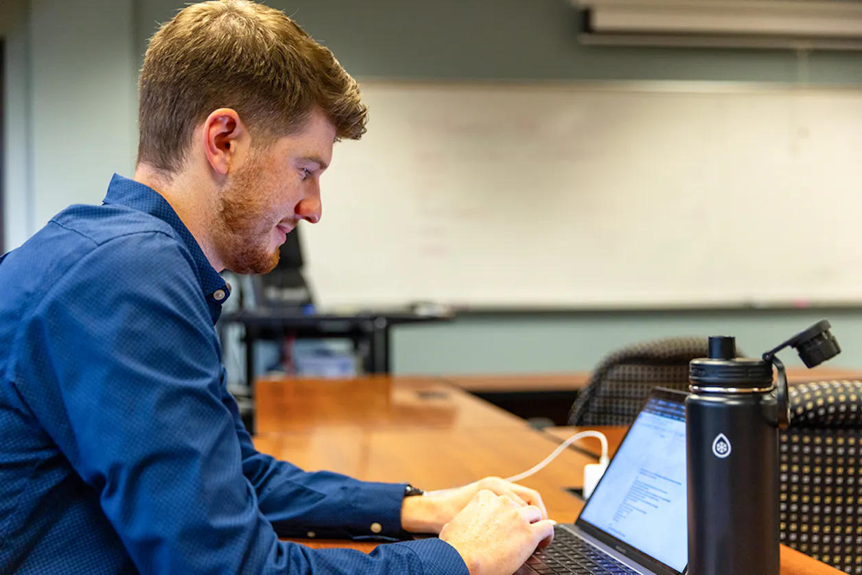 A graduate student works intently on a laptop in a university classroom, focused on an assignment with a water bottle and notebooks nearby.