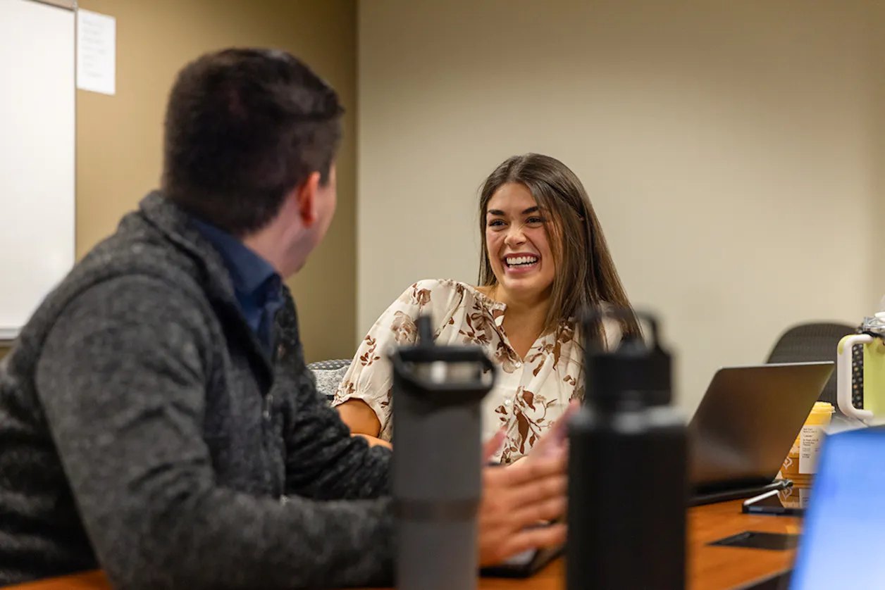 A smiling college student talks with a peer in a classroom setting, both working on laptops during a collaborative learning session.