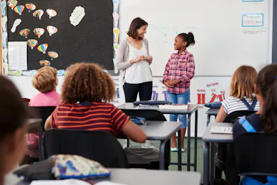 An elementary teacher and a student stand at the front of the class, sharing a moment while other students look on during a classroom presentation.