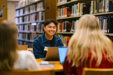 Smiling student at a library table with laptop and books, chatting with classmates.