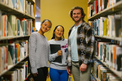 Three smiling students standing between library bookshelves at Bethel University.