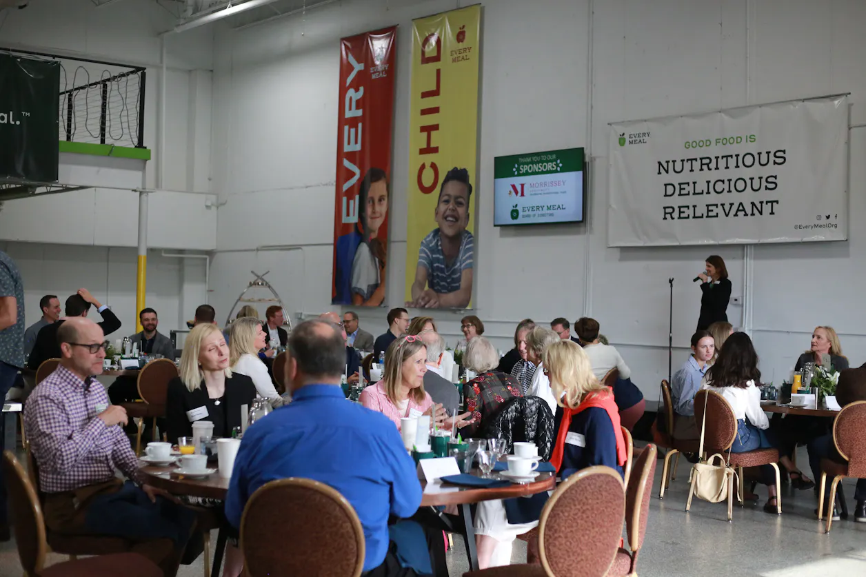 Every Meal Board Chair Jenna Soule ’95 stands on a stage in front of a crowd sitting at tables in Every Meal’s warehouse.