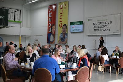 Every Meal Board Chair Jenna Soule ’95 stands on a stage in front of a crowd sitting at tables in Every Meal’s warehouse.