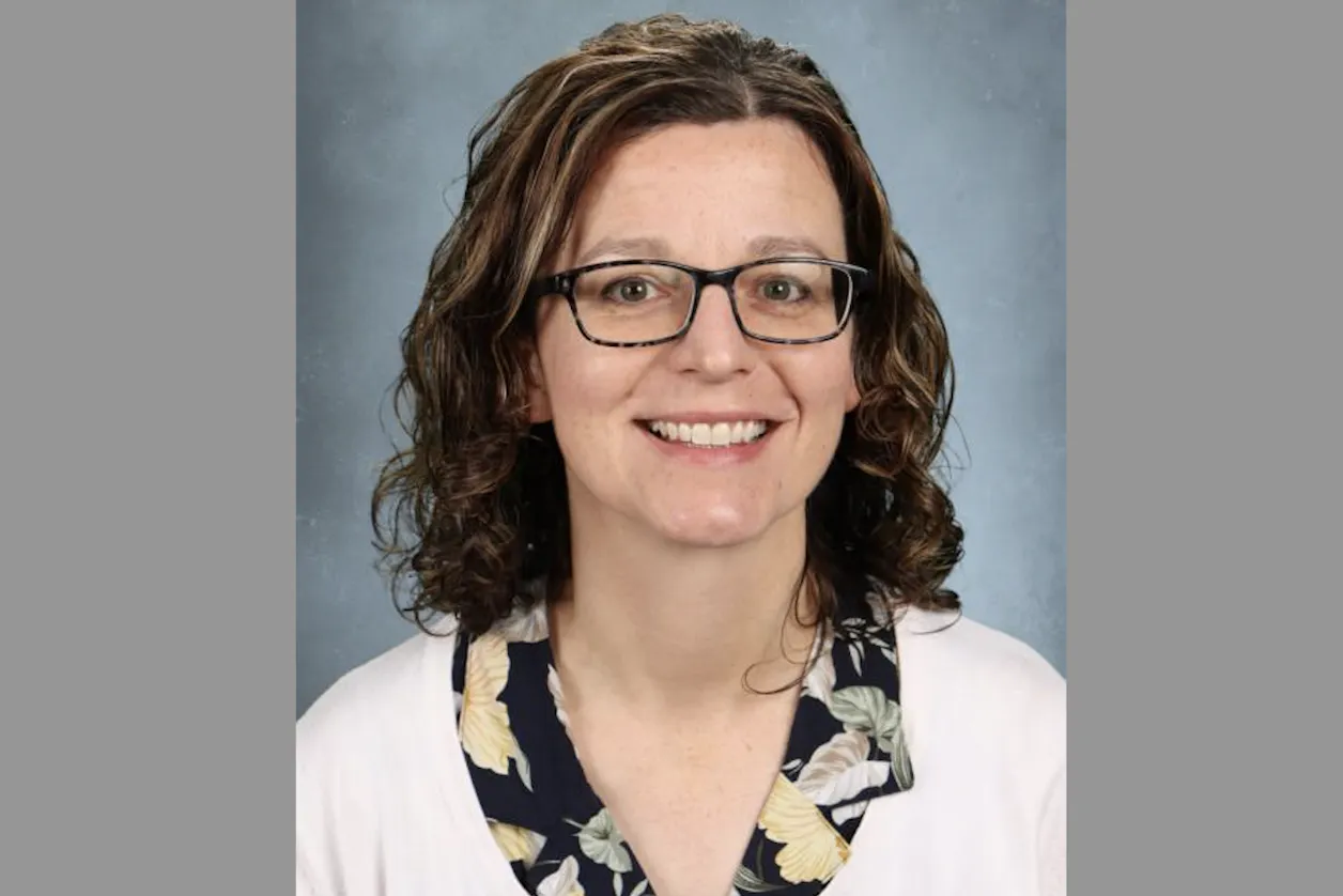 Smiling woman with curly brown hair, wearing glasses and a floral blouse with a light cardigan.