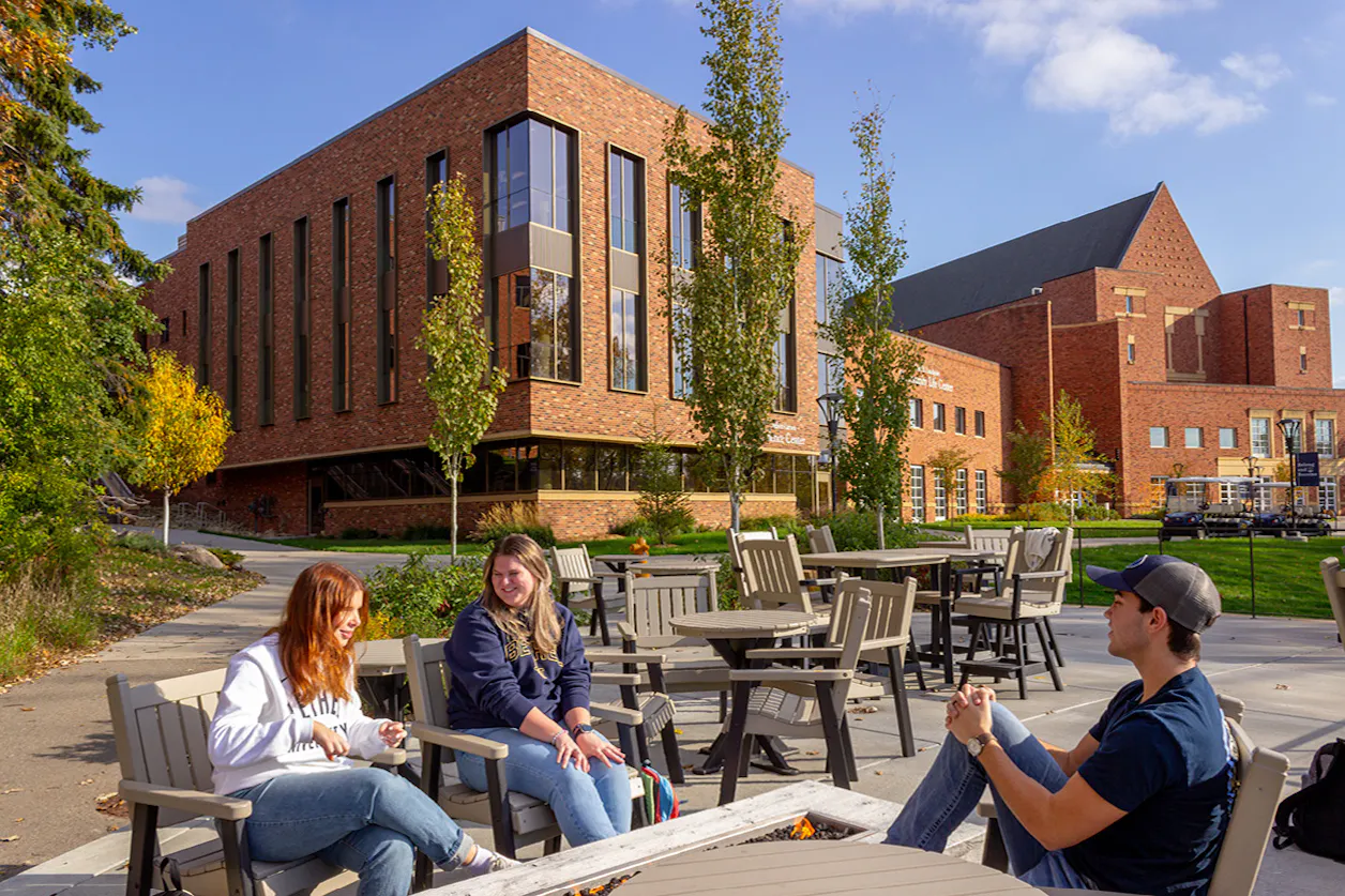 Three BEthel students char while sitting around a fire pit before Bethel's brick building with leaves showing the first signs of changing colors. 