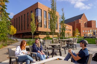 Three BEthel students char while sitting around a fire pit before Bethel's brick building with leaves showing the first signs of changing colors. 