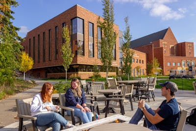 Three BEthel students char while sitting around a fire pit before Bethel's brick building with leaves showing the first signs of changing colors. 