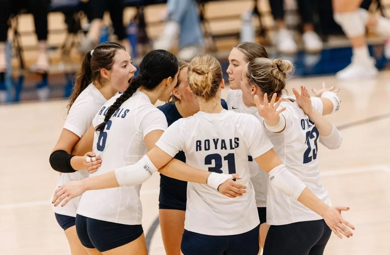 Bethel volleyball players huddle on the court, arms linked in a moment of team unity.