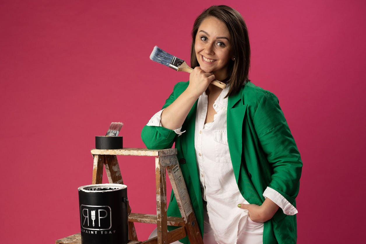 BIllie Asmus, "the RePaint Lady," poses on a ladder with painting supplies, before a hot pink background