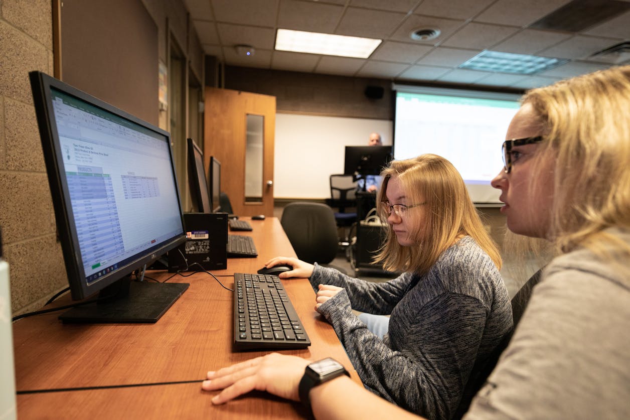 Two students use desktop computers together in a computer lab setting.