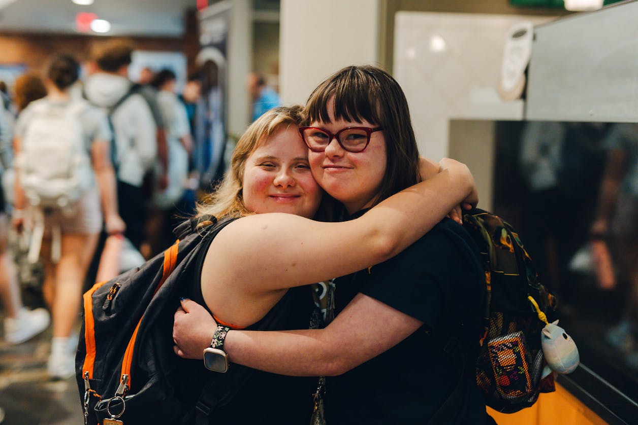 Two students hug and smile in a crowded campus space.