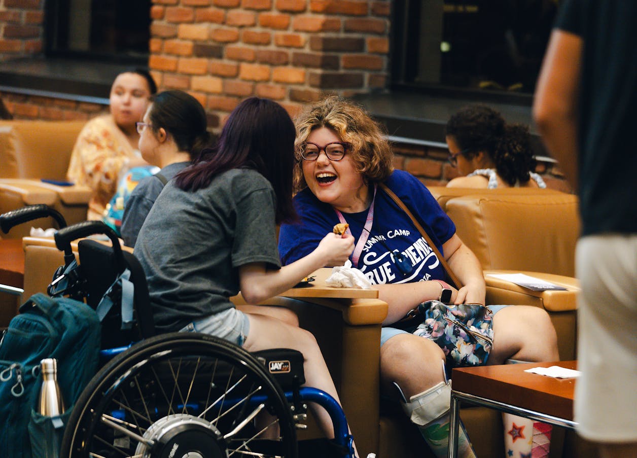 Two students in wheelchairs laugh and share food in a lounge area.