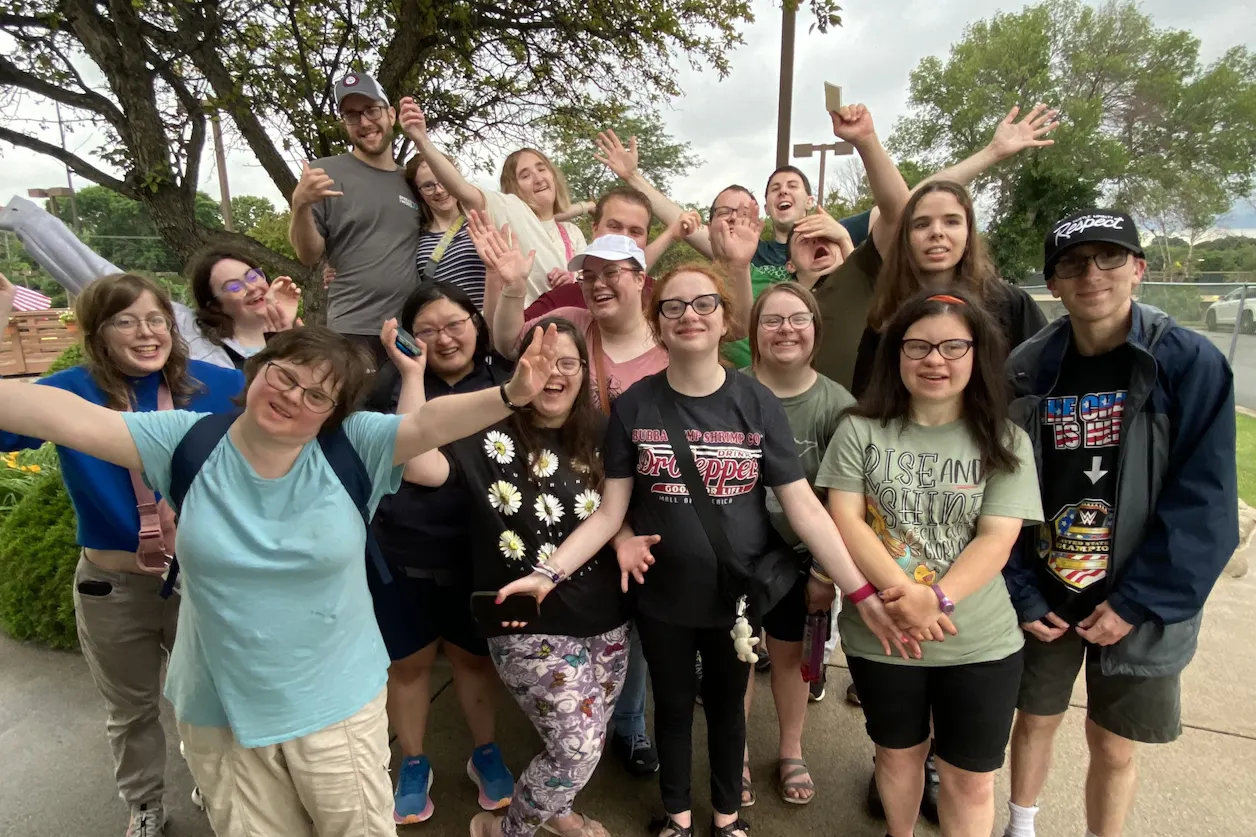 A large group of smiling BUILD alumni and friends pose together outdoors on a sidewalk, some raising their arms playfully. Trees and green space are visible in the background on a cloudy day.