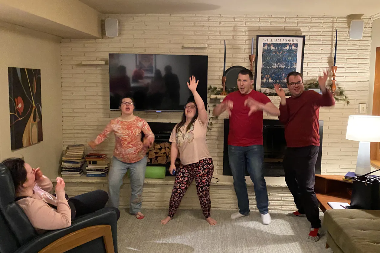 Group of friends dancing and laughing in a cozy living room while others watch and smile.