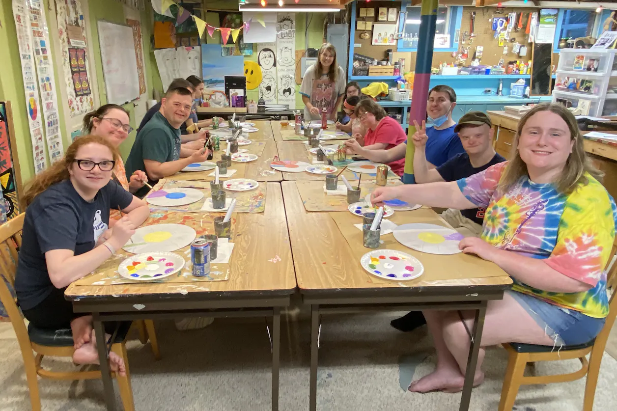 Group of people painting colorful art at a long table in a cheerful, creative studio space.