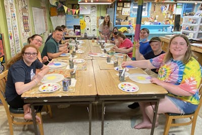 Group of people painting colorful art at a long table in a cheerful, creative studio space.