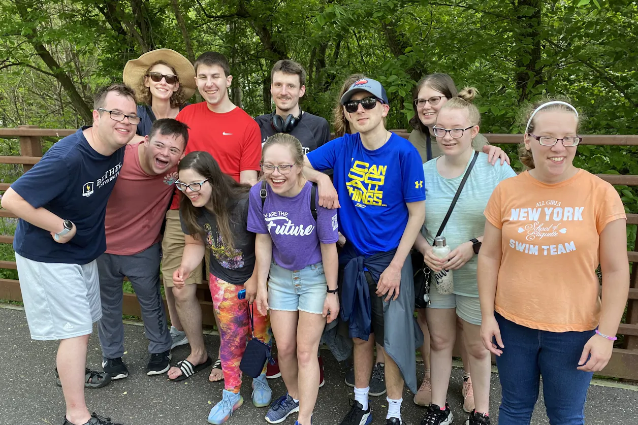 Group of friends smiling together on a forest trail near a railing during a sunny day outdoors.