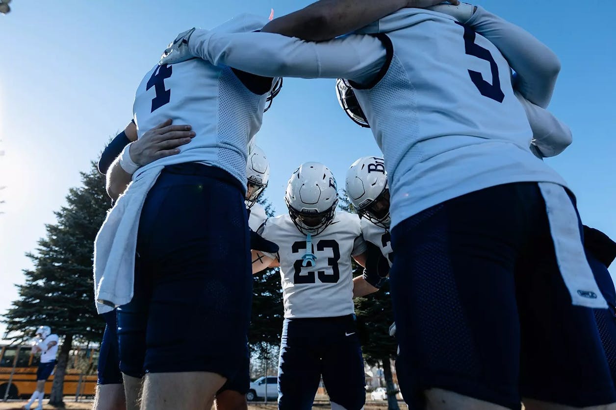 Bethel University football players stand in a tight pregame huddle on the field, arms around each other under a clear blue sky.