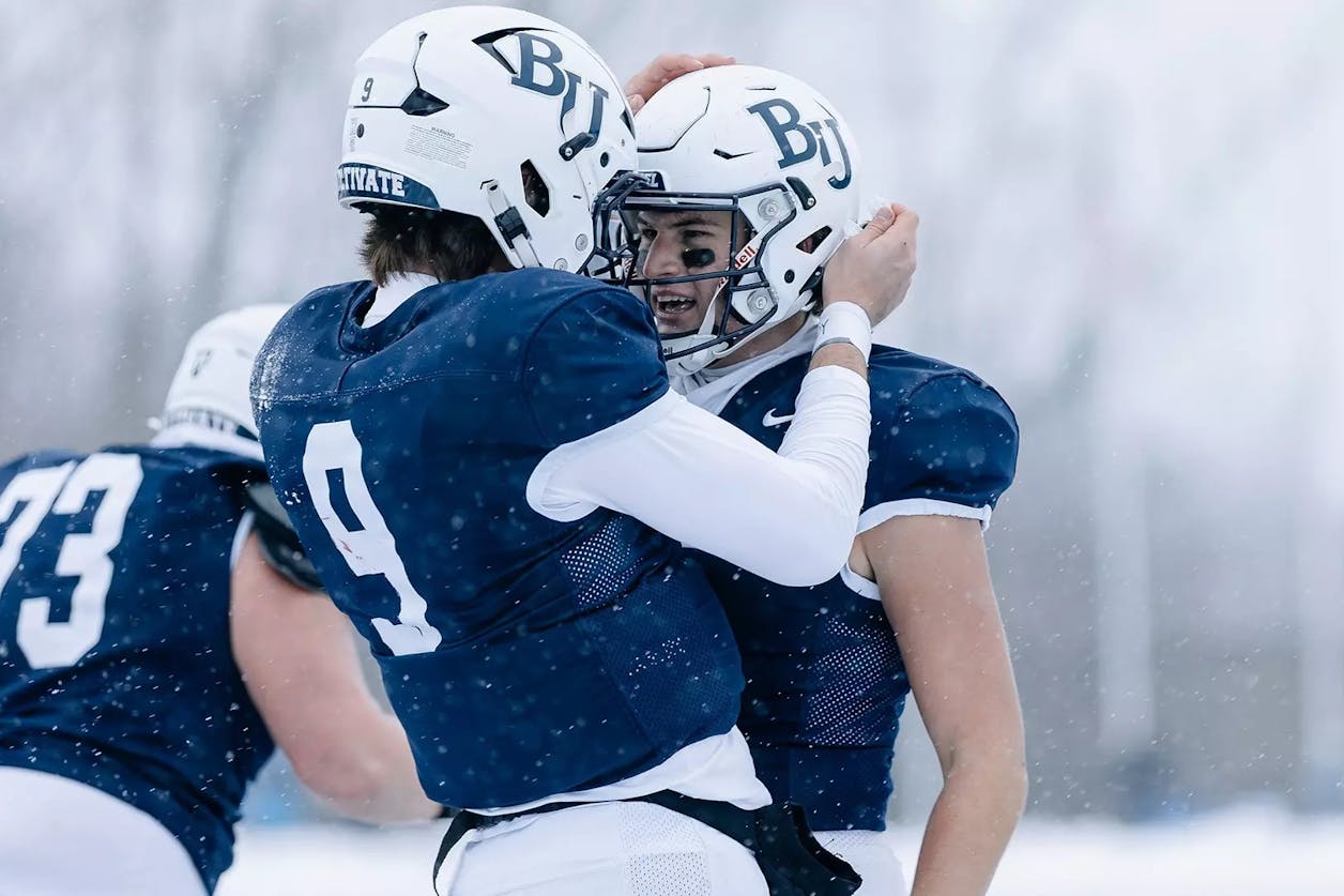 Two Bethel University football players celebrate on the field during a snowy game, wearing navy jerseys and white helmets. 