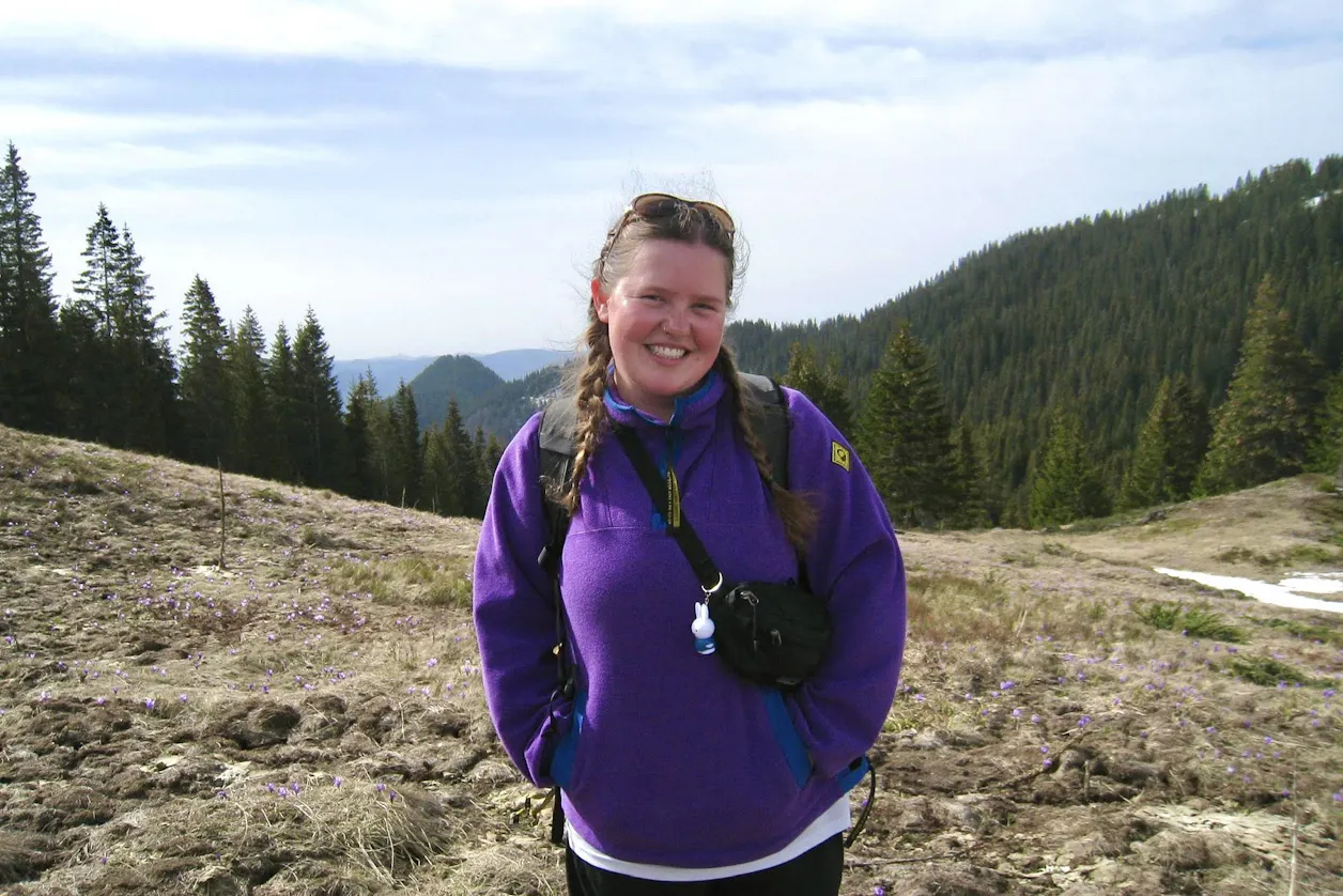 Smiling woman in a purple jacket with braids stands in a mountain meadow with pine trees.