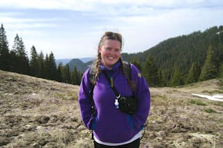Smiling woman in a purple jacket with braids stands in a mountain meadow with pine trees.