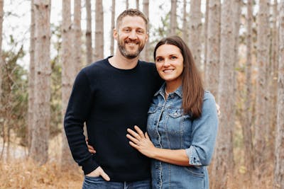 Kevin Pieh and Kayla Pieh, both Bethel alumni, smile together in a wooded setting. Kevin, CEO of Ellsworth Cooperative Creamery, wears a navy sweater, while Kayla, in a denim jacket, rests her hand on his chest.