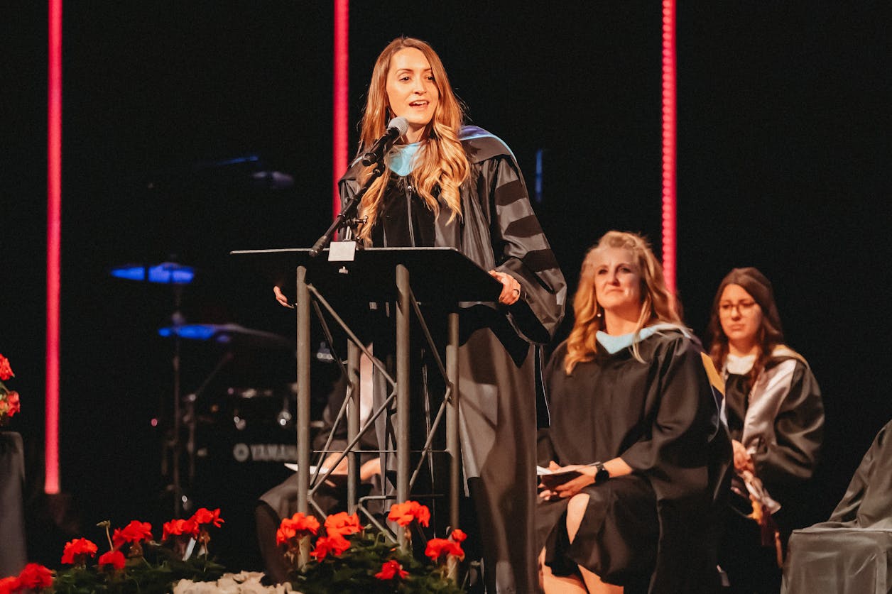 Shawn Lohse stands at a podium in academic regalia, delivering a commencement speech.