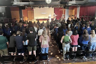 A large group of Royal Academy students stands in rows during Chapel, facing a stage where two worship leaders lead a song with lyrics projected on the screen behind them.
