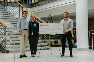 Three individuals pose next to a large $37,000 grant check from State Farm to Bethel University Royal Academy, displayed on an easel in the Brushaber Commons.