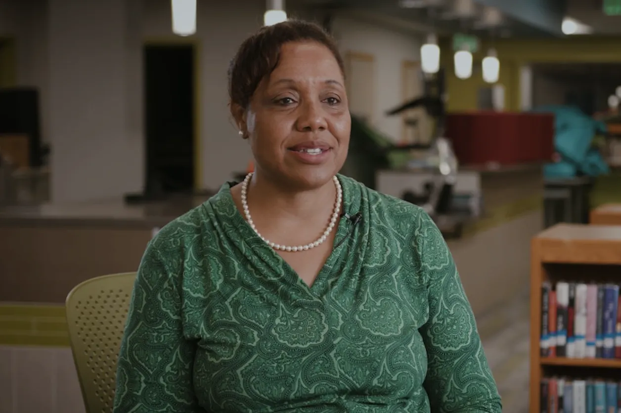 Stacie Stanley in a green patterned top and pearls speaks in a well-lit library or study space.