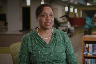 Stacie Stanley in a green patterned top and pearls speaks in a well-lit library or study space.