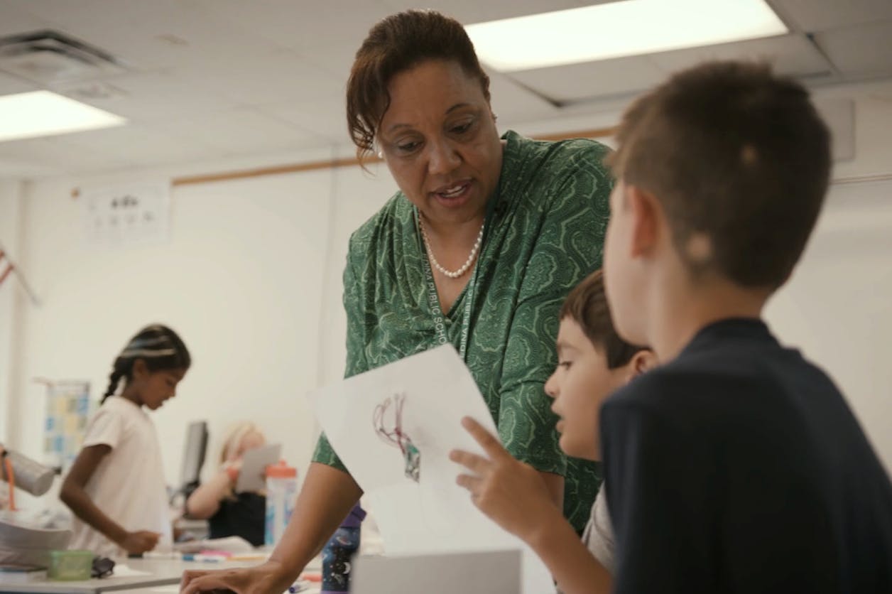 Educator in green helps two young students review a drawing during classroom activity time.
