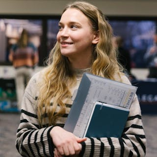 Student in a striped sweater smiles while holding folders labeled Bethel Advance at an event.