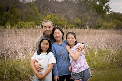 Tao Wu with his wife, Jing Fang, and daughters Elisha and Yvonne, smiling outdoors in a grassy field.