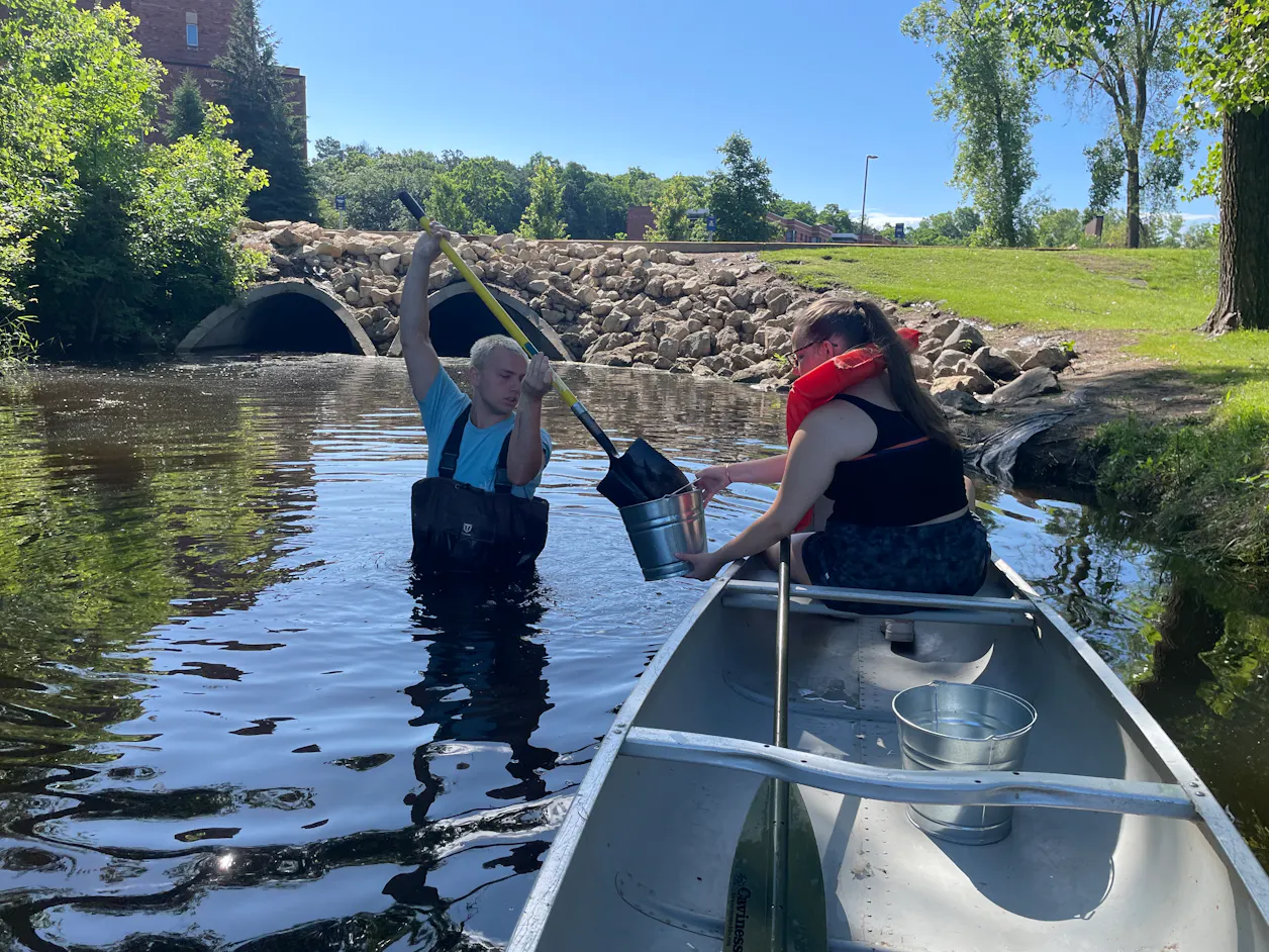 Alex Michuda ’26 stands in Lake Valentine, handing a sediment sample to Karissa Nelson ’27 in a canoe.