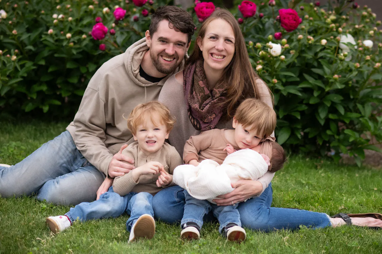 Greg Stewart poses with his wife and three children in the grass with large, bright pink flowered bushes behind them. 