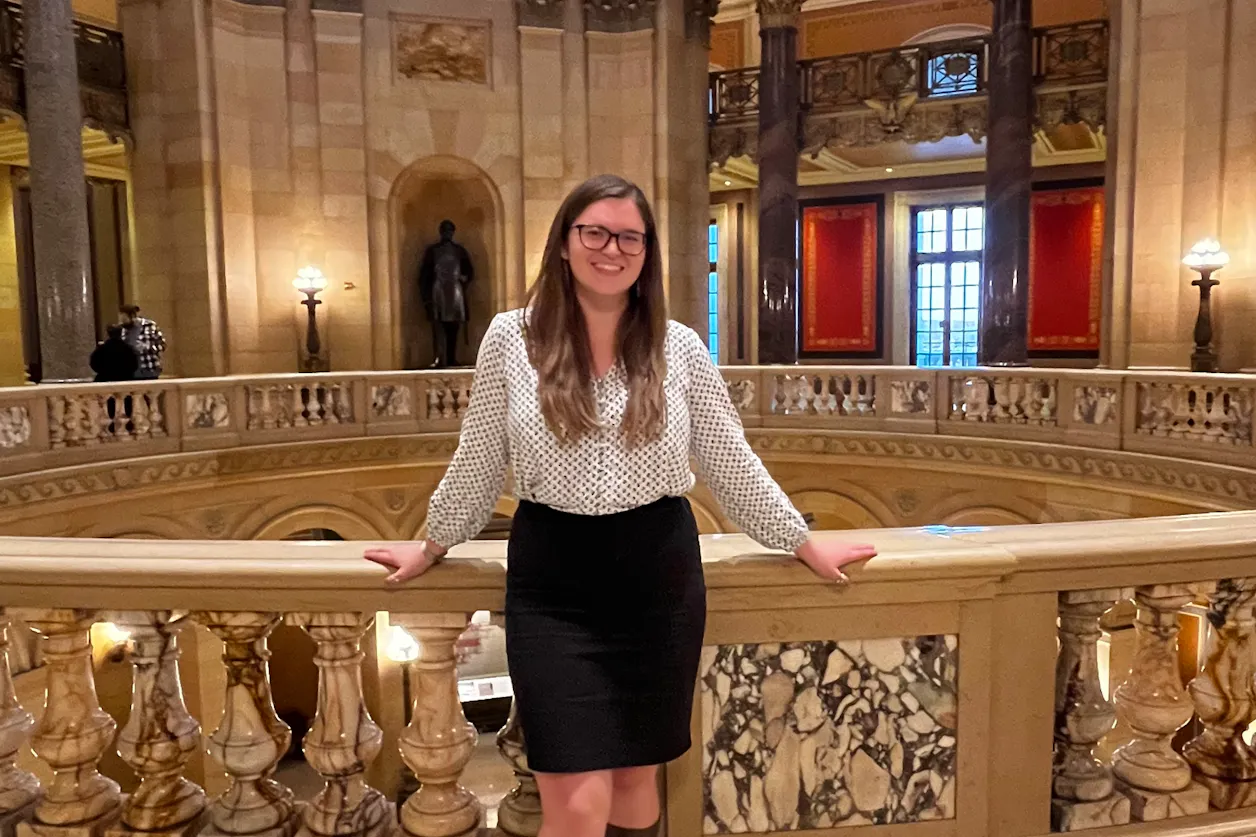 Woman wearing glasses smiles while standing along a marble balcony inside the Minnesota State Capitol.