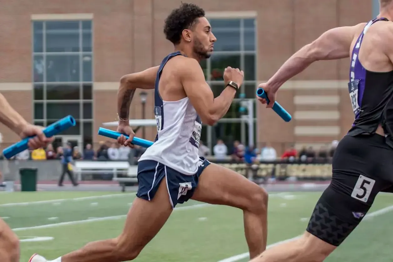 Bethel men's track athlete sprints in a relay race while carrying a blue baton.