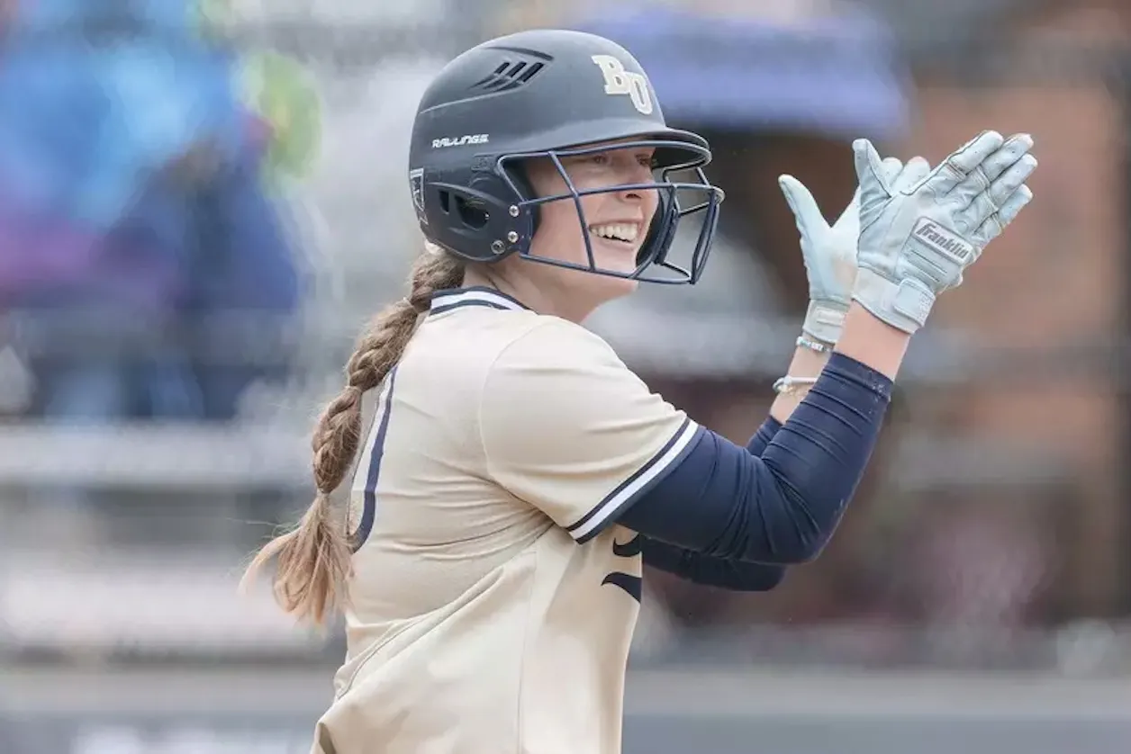 Bethel softball player in a batting helmet smiles and applauds from the field.