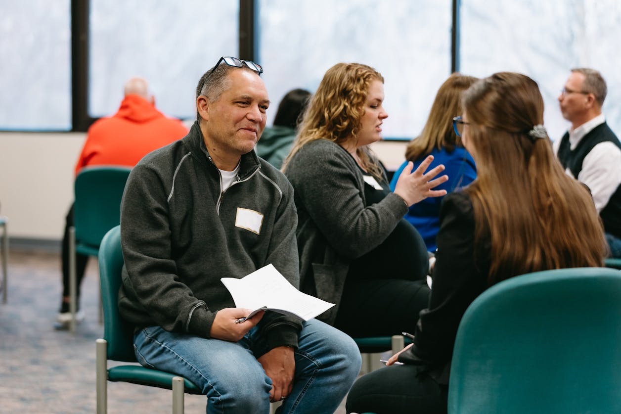 Two adults sit facing each other during a small group discussion at a professional workshop, holding papers while other participants talk in the background.