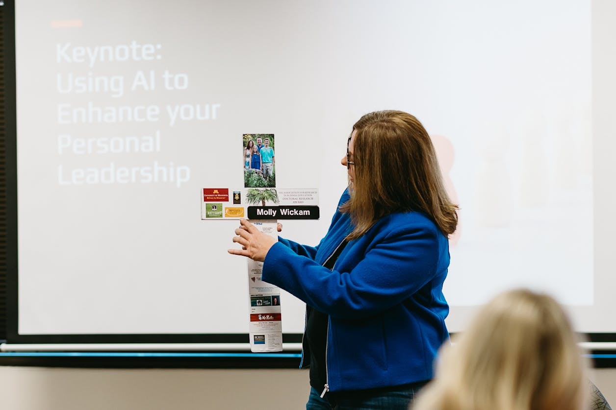 Molly Wickam stands in front of a presentation screen holding a cross-shaped display with photos and mementos as she introduces herself during a keynote presentation about using AI in leadership and education.