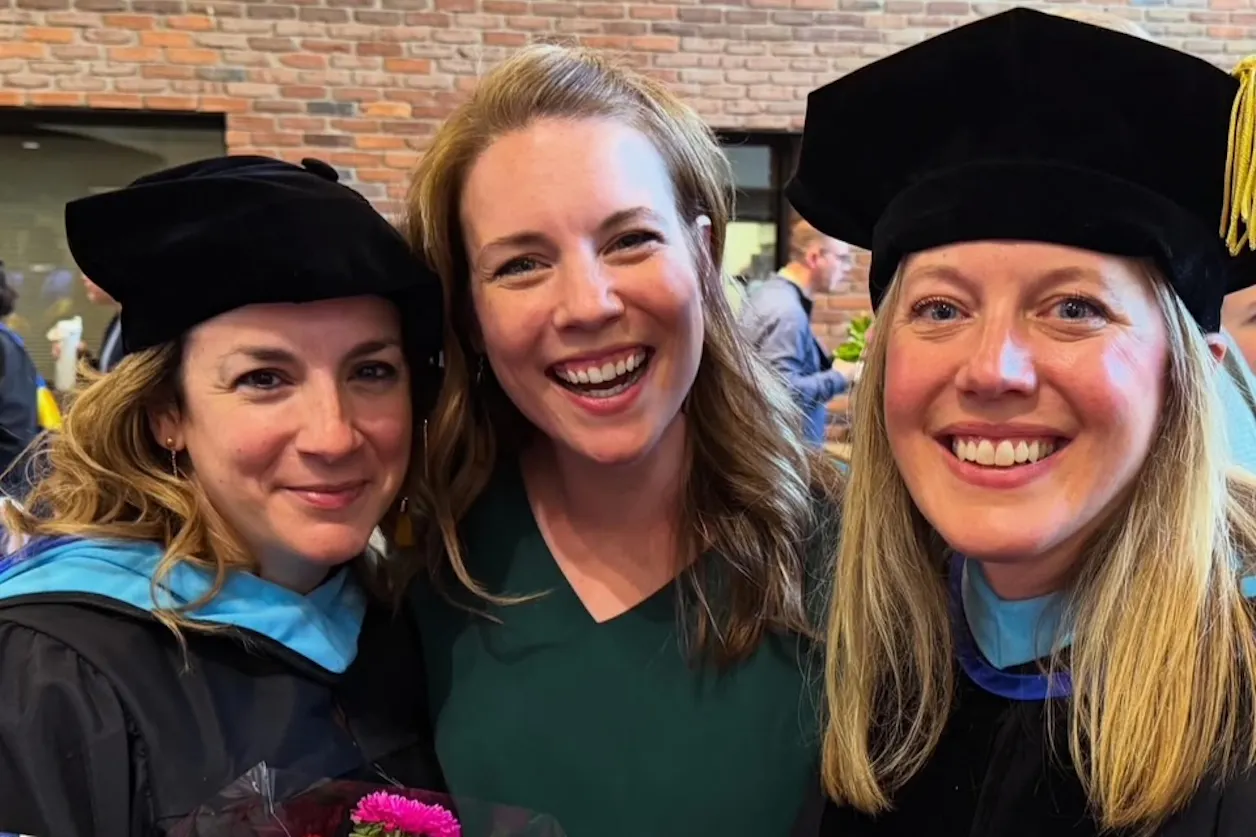 Three women smile for the camera, two in doctorate graduate regalia and the other in plain clothes. 